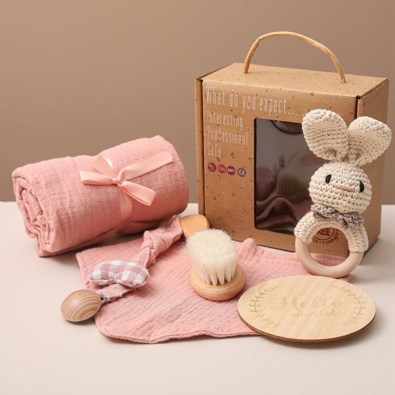 Das Amelia Baby Geschenk zur Geburt von Hello World besteht aus einer rosa Decke, einer hölzernen Haarbürste, einem Rasselhase, einem rosa-weiß karierten Beißring, einer Meilenstein-Fototafel und einer hölzernen Rassel - alles schön arrangiert in einer braunen Box.