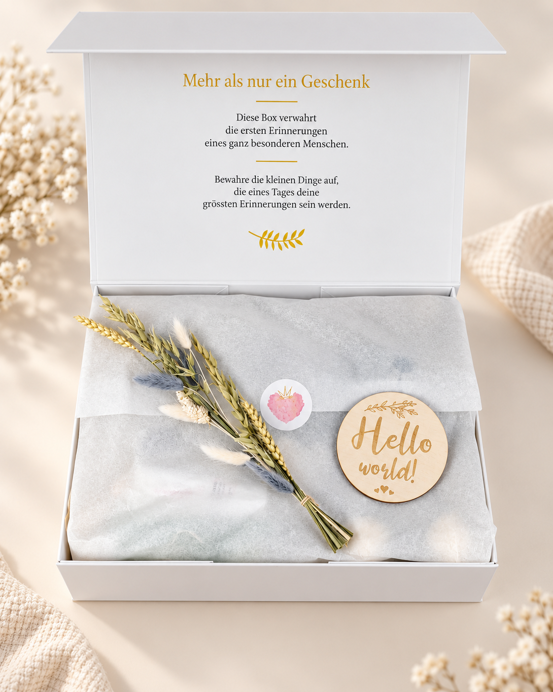 Geöffnete cremeweisse Premium Geschenkbox mit Erinnerungsaufdruck im Innendeckel, Seidenpapier, Trockenblumen und Holzanhänger „Hello world!“ auf hellem Hintergrund.