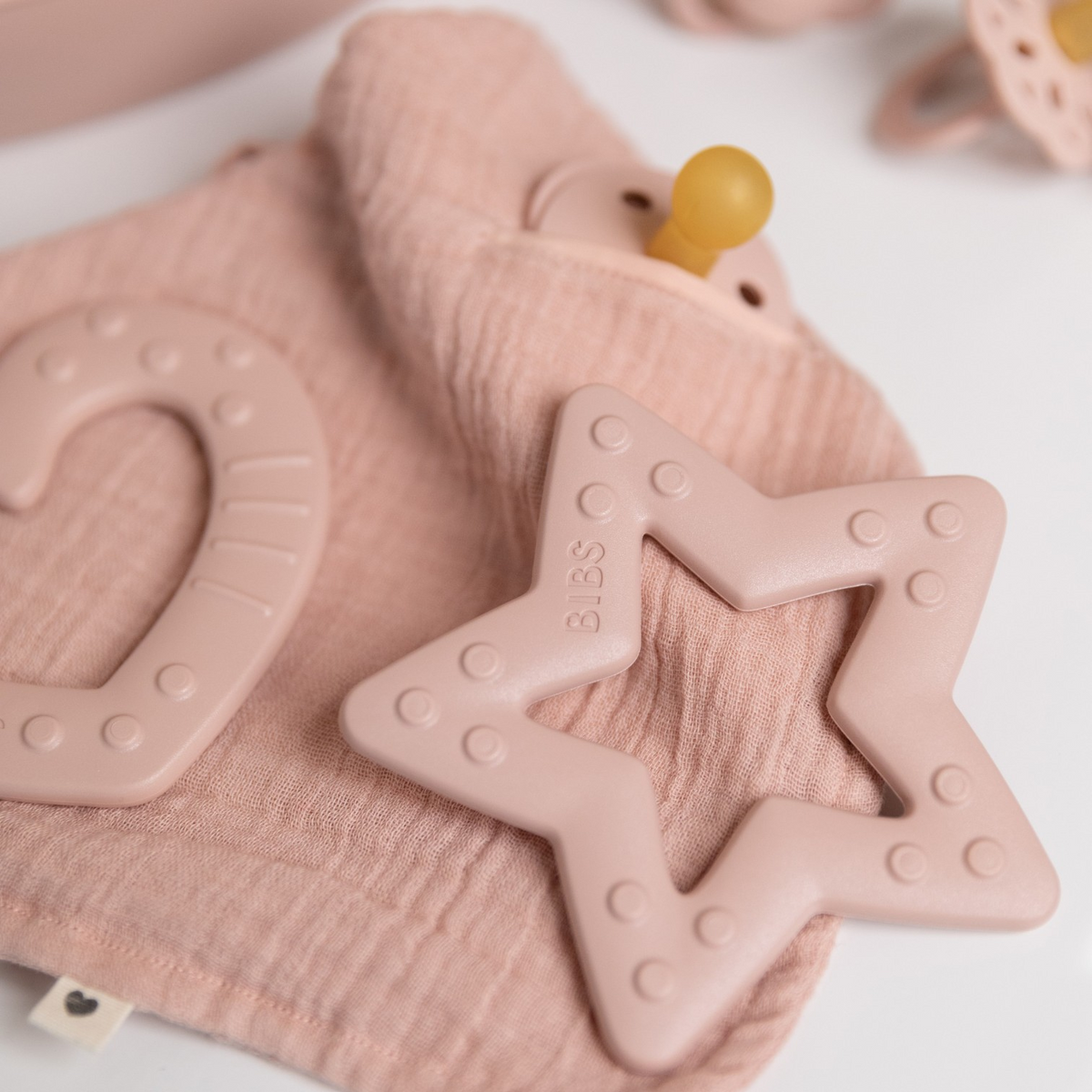 Star-shaped teething ring in rosé as a teething aid for babies on a gauze cloth next to a pacifier and heart teething ring