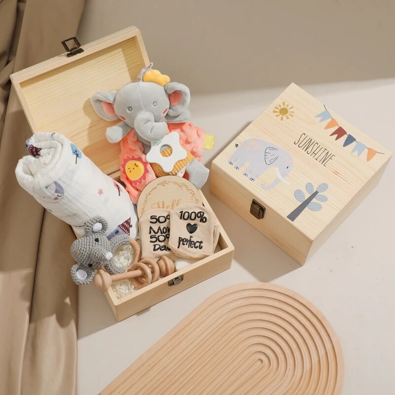 Die "Cyrill Baby Geschenk zur Geburt" Holzbox von Hello World steht samt Plüschelefant, Decke, Holzplättchen, gehäkelter Rassel und Schnuller sowie verziertem Deckel auf einer beigen Fläche.