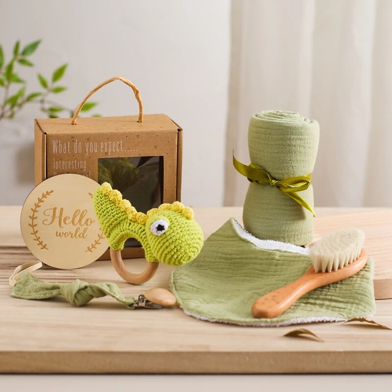 Das Hello World Alfredo Baby Geschenk zur Geburt Set besteht aus einem grünen gehäkelten Dinosaurierring, einer Holzbürste, einer Schnullerkette, einem grünen Mulltuch, einem aufgerollten Wickeltuch mit Schleife und einem hölzernen "Hello World"-Schild auf der Vorderseite der Box.