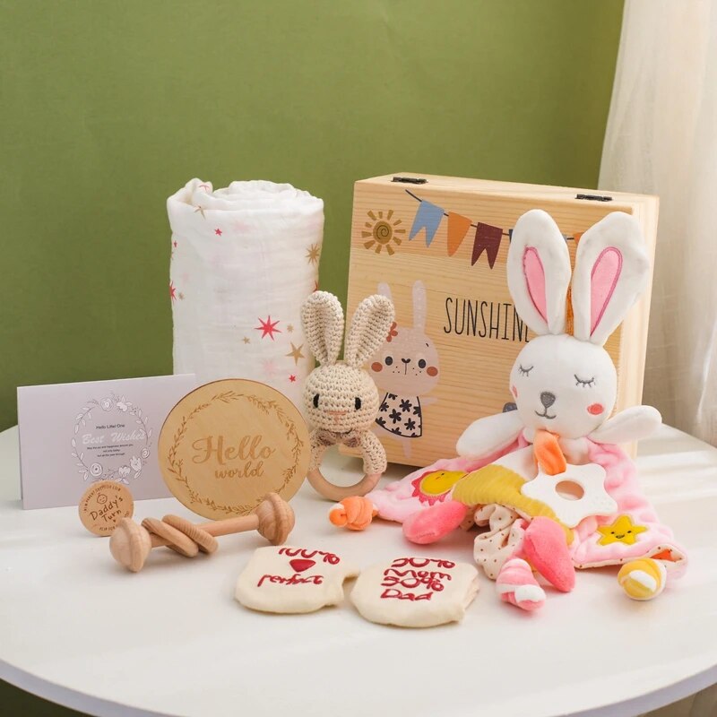 Das Anna Baby Geschenk zur Geburt von Hello World besteht aus einer Sunshine-Holzbox, Plüsch-Rasselhasen, einer Decke mit rosa Sternen, Holzrasseln und -spielzeug, Meilensteinkarten und Babysocken - perfekt präsentiert auf einer hellen Oberfläche mit grünem Hintergrund.