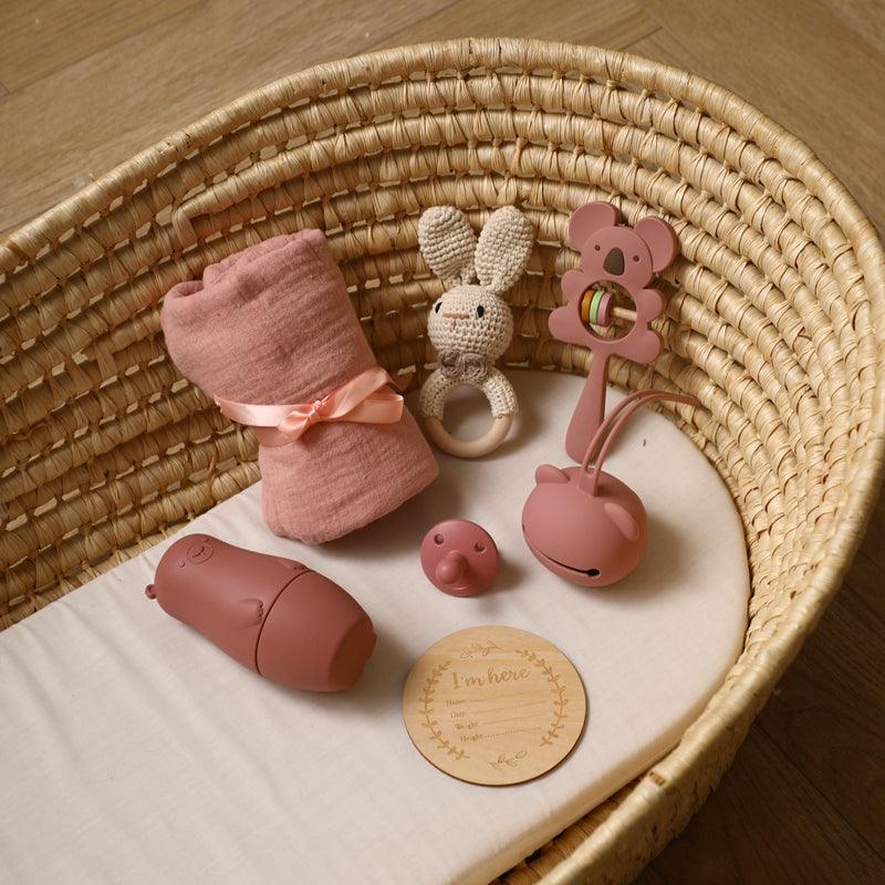 Das "Ayla Baby Geschenk zur Geburt" von Hello World besteht aus einem gewebten Stubenwagen mit rosa Babydecke, gehäkelter Hasenrassel, Koala-Greifling, Silikon-Schnuller, Holzplakette und rosa Schnullertasche auf einer weißen Matratze.