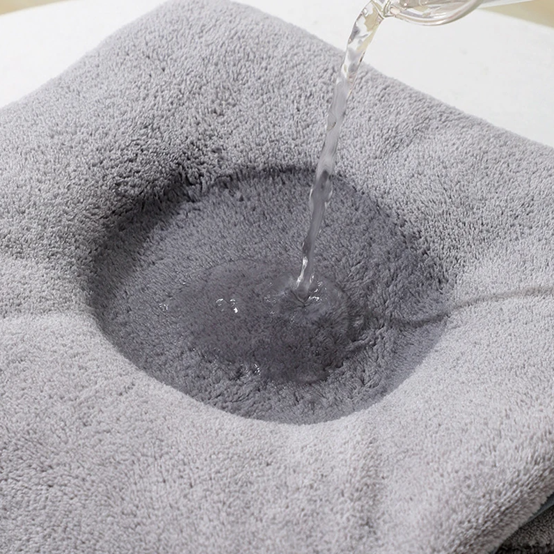 Ein Wasserstrahl ergießt sich auf ein hellgraues Hello World Badetuch mit Kapuze Elefant (80x80cm) und erzeugt einen dunklen, nassen Fleck in der Mitte des flauschigen Handtuchs.