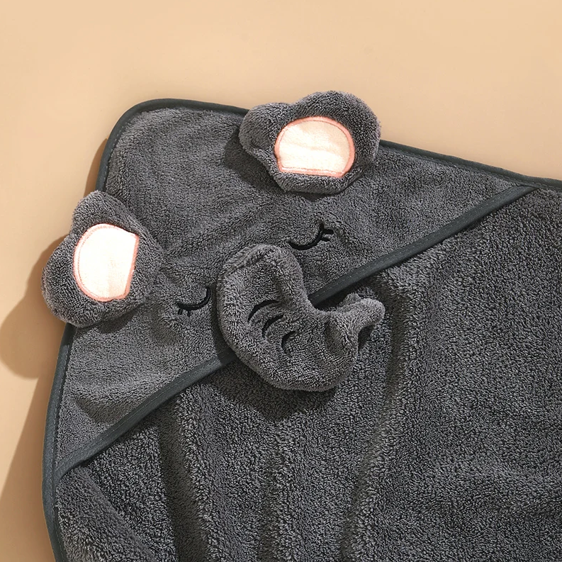 Ein weiches, dunkelgraues Hello World Badetuch mit Kapuze Elefant (80x80cm) in Elefantenform mit Kapuze, Stoffohren, Rüssel und aufgenähten Augen wird auf einer neutralen beigen Fläche präsentiert.