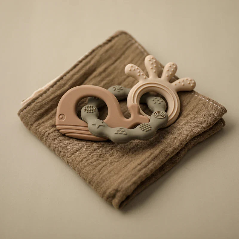 Ein grüner walförmiger Silikonbeißring von Hello World (Beissring Walfisch Grün, 10,5x7cm) ruht auf einem gefalteten braunen Tuch vor einem neutralen Hintergrund.