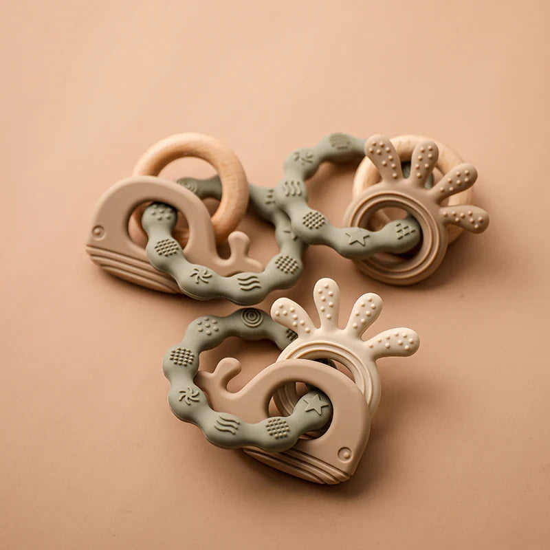 Zwei Hello World Beissring Walfisch Grün Baby-Zahnspielzeug (10,5x7cm) mit walförmigen Silikonkörpern, strukturierten Ringen und Holzgriffen in zartem Grün und Creme, auf beigefarbenem Hintergrund.