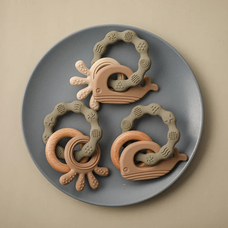 Ein Hello World Beissring Walfisch Grün Beißspielzeug (10,5x7cm) mit einer strukturierten Oberfläche in Grün wird auf einem runden grauen Teller vor einem hellbeigen Hintergrund präsentiert.