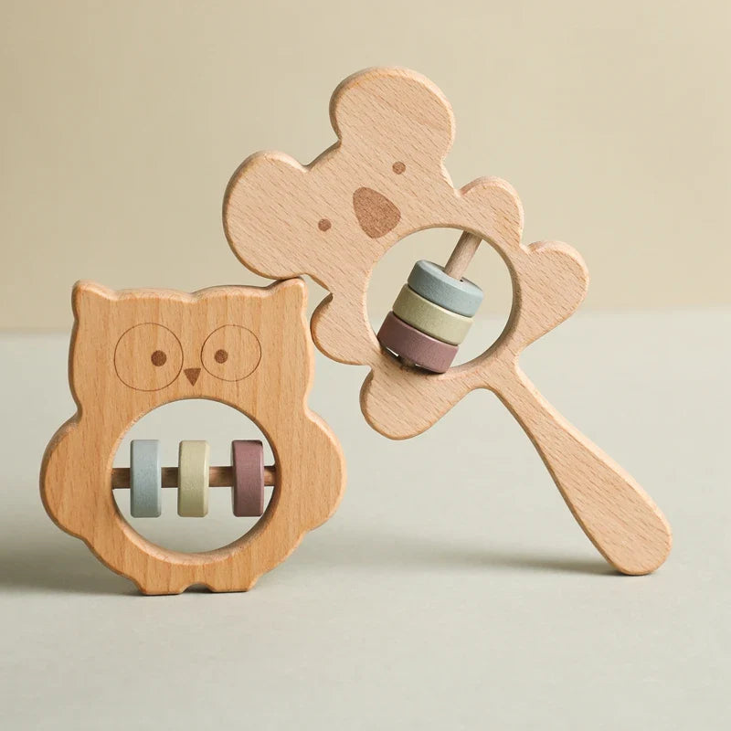 Zwei nachhaltige Babyrasseln aus Holz von Hello World, darunter die 8x8cm große "Eule aus Holz" und ein Bär, jeweils mit pastellfarbenen Ringen in Ausschnitten. Beide ruhen auf einer beigen Oberfläche vor einem neutralen Hintergrund.