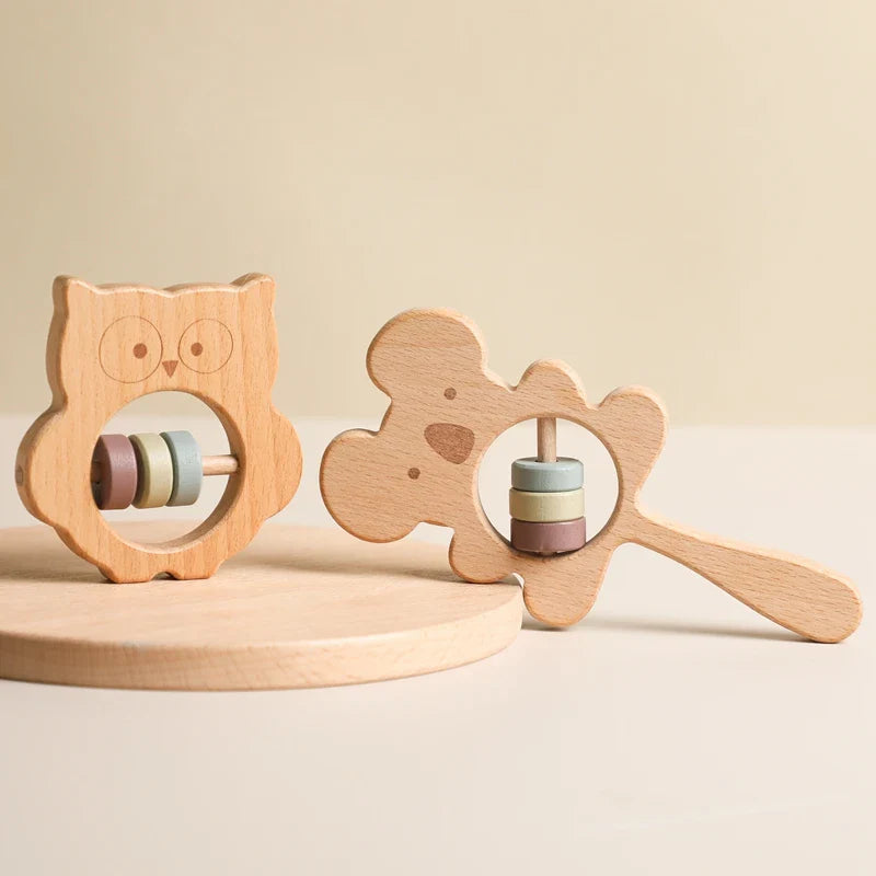 Zwei hölzerne Babyrasseln, darunter die bezaubernde Hello World Eule aus Holz (8x8cm) und ein Bär mit pastellfarbenen Ringen, werden auf einem hellen, runden Holzsockel präsentiert - das perfekte Naturholz Spielzeug für jedes Eule Kinderzimmer.