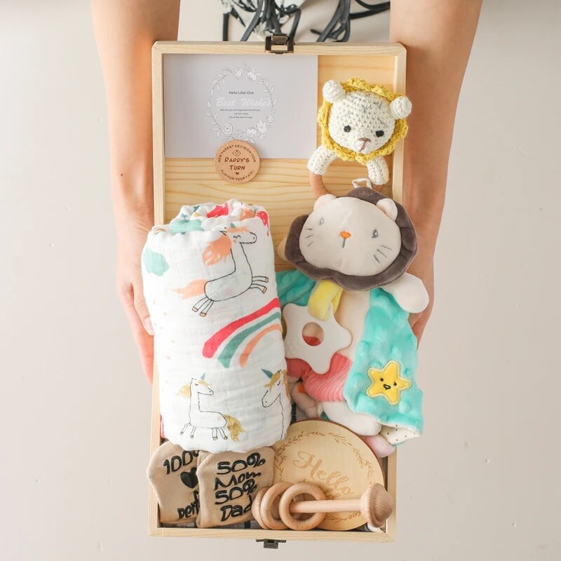 Eine Person hält eine geöffnete nachhaltige Holzbox von Hello World's Jannis Baby Geschenk zur Geburt, gefüllt mit Babyartikeln wie einer Einhorndecke, Rassellöwe, Bärenplüschtieren, Holzschnuller, Schnuller, Lätzchen und einer runden Holzplatte.