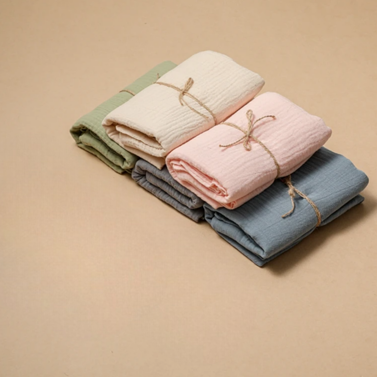 Vier handgefertigte Musselintücher von Hello World in Grün, Creme, Rosa und Blau-je 55x65cm-werden mit einer Kordel gebunden und auf hellem Beige arrangiert, als stilvolle Alternative zum Multifunktionstuch Grau.