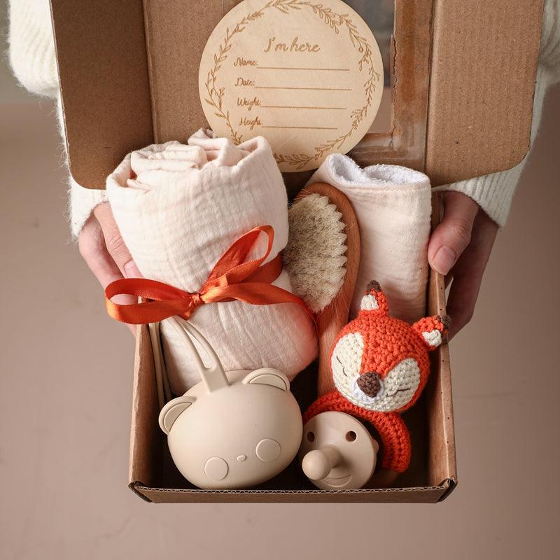 Eine Person hält ein geöffnetes Rico Baby Geschenk zur Geburt von Hello World in der Hand, das eine gerollte Decke, eine weiche Bürste, eine Karte, ein Schnuller-Set, einen Rasselfuchs und einen Einhorn-Beißring enthält - alles schön mit einer Schleife arrangiert.