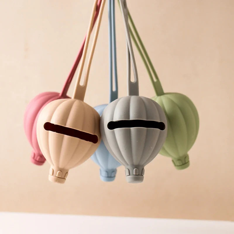 Vier Hello World Schnullertasche Heissluftballon Grau (7x22,5cm) Schlüsselanhänger in Pastellrosa, Beige, Blau und Grün, jeweils mit einem Frontschlitz für Schnuller Aufbewahrung, hängen nebeneinander vor einem einfarbig beigen Hintergrund.