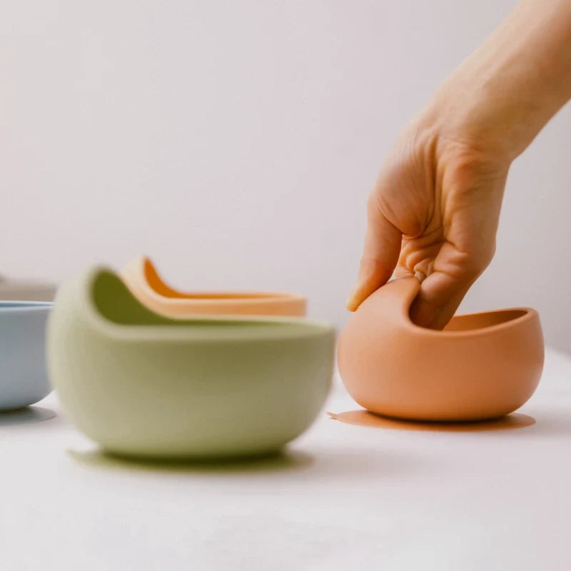 Eine Hand greift nach einer grauen Hello World Schüssel rund (12x8cm), BPA-frei, mit gewölbter Öffnung - ideal für Baby-Led Weaning - neben ähnlichen Schüsseln in Grün und Blau auf einer weißen Oberfläche.