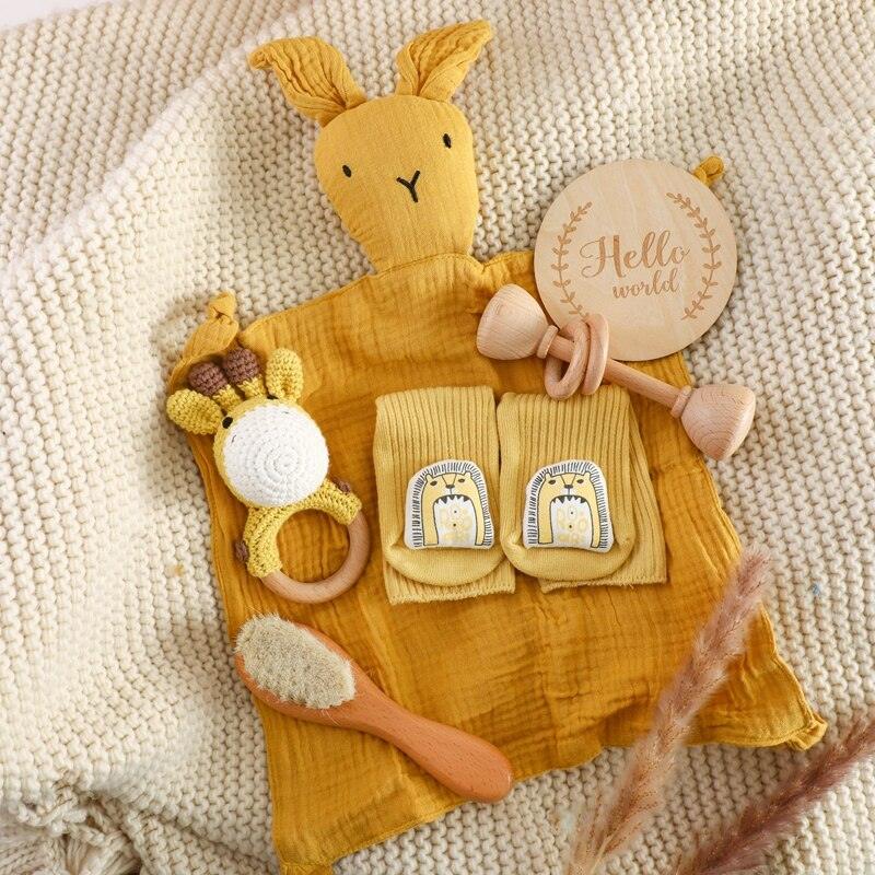 Auf einer beigen Strickdecke liegt das Shania Baby Geschenk zur Geburt von Hello World - komplett mit gelber Hasendecke, Rasselgiraffe, niedlichen Söckchen mit Bärengesicht und einem Hello World Holzschild. Das ideale nachhaltige Babygeschenk!.