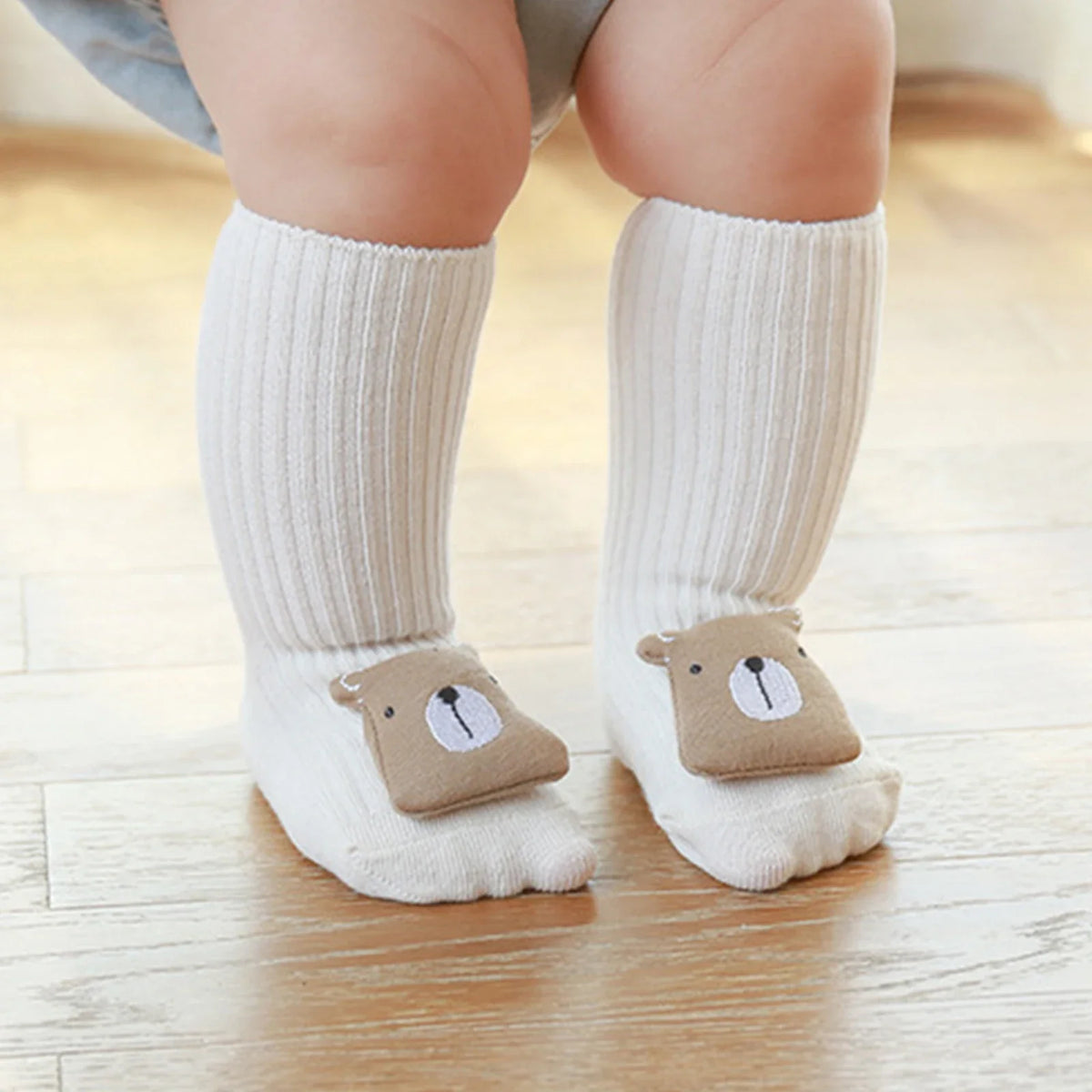 Ein Baby, das Hello World Socken Bär (Sohle: 6x11cm, 6-12 Monate) mit einem niedlichen beigen Bärendesign trägt, sitzt auf einem hellen Holzboden.