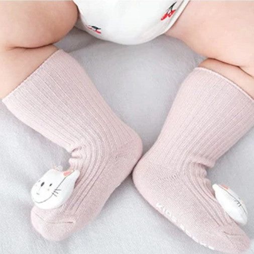 Ein Baby mit pummeligen Beinen trägt pale pinke Hello World Socken Katze (6-12 Monate, Sohle 6x11cm) mit weißen Katzen auf den Zehen und sitzt auf einer hellen Oberfläche.