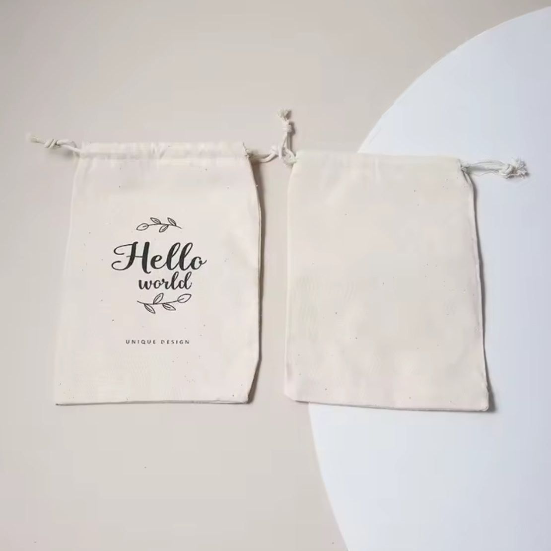 Zwei beigefarbene Spielzeugbeutel "Hello World" (14,5 x 23,5 cm) von Hello World liegen nebeneinander: der eine zeigt das Hello World-Design mit Akzenten, der andere ist einfarbig - ein charmantes Geschenkset zur Geburt eines Babys.