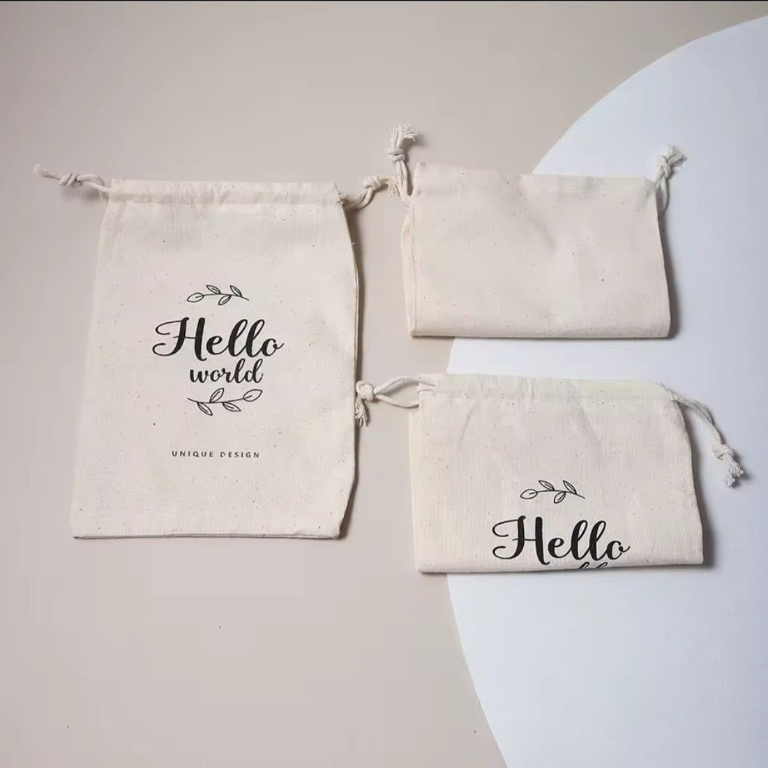 Drei cremefarbene Baumwollbeutel von Hello World ruhen auf einem hellen Hintergrund. Auf dem größten Spielzeugbeutel "Hello World" (14,5 x 23,5 cm) steht "Hello world", auf den beiden kleineren "Hello", alle mit minimalem Blattdesign - ideal als Geschenk zur Geburt.