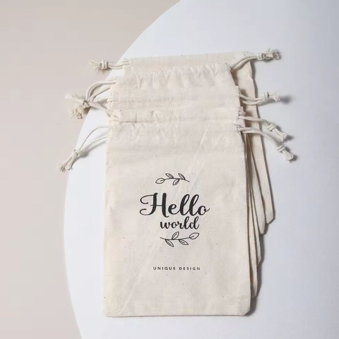 Ein Stapel beigefarbener Spielzeugbeutel "Hello World" (14,5x23,5cm) von Hello World liegt auf einer weißen Fläche; der oberste Beutel zeigt dekorative Blätter und UNIQUE DESIGN - ideal als Geschenk zur Geburt.