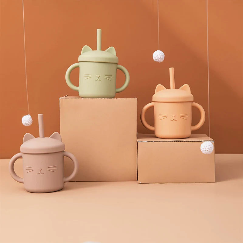 Drei beigefarbene Hello World Trinkbecher Katze (12x11cm) mit Katzengesicht und Griffen sind auf Pappschachteln vor einem warmen braunen Hintergrund ausgestellt, darüber hängen weiße Kugeln an Schnüren.