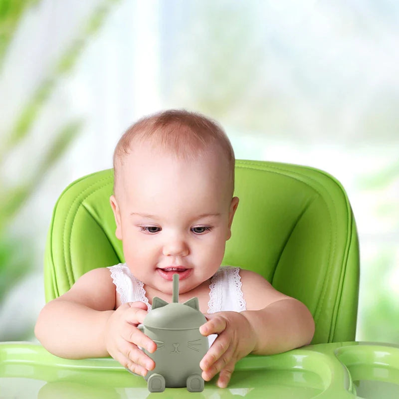 Ein Baby sitzt in einem grünen Hochstuhl und schaut neugierig auf einen Hello World Trinkbecher Katze sitzend Grün (13x11cm) mit Strohhalm. Der Becher hat die Form einer Katze, und der sanft verschwommene Hintergrund zeigt grüne und weiße Töne.