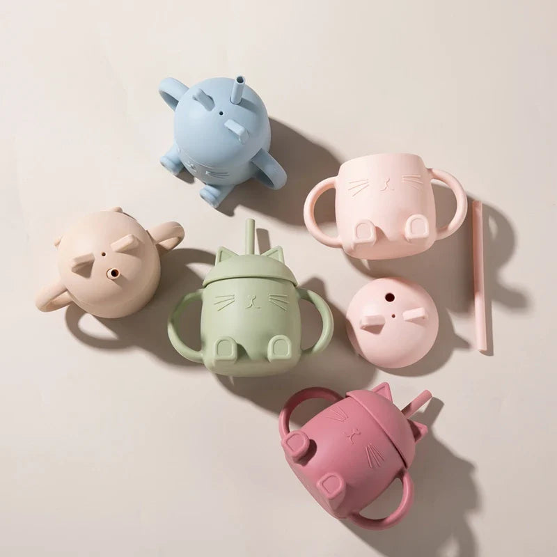 Der grüne Trinkbecher "Katze sitzend" von Hello World (13x11cm) mit Henkeln und Deckel steht auf einer hellen Fläche, daneben liegt ein rosa Strohhalm. Das Design ist naturverbunden und freundlich gestaltet.