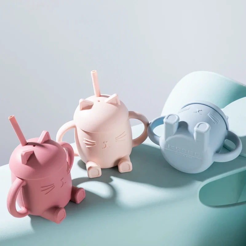 Drei pastellfarbene Trinkbecher - ein blauer Becher in Bärenform und zwei Hello World Trinkbecher Katze sitzend Pink (13x11cm) - stehen auf einer weichen hellblauen Unterlage. Jeder Becher hat einen Strohhalm und kleine Griffe zum einfachen Greifen.