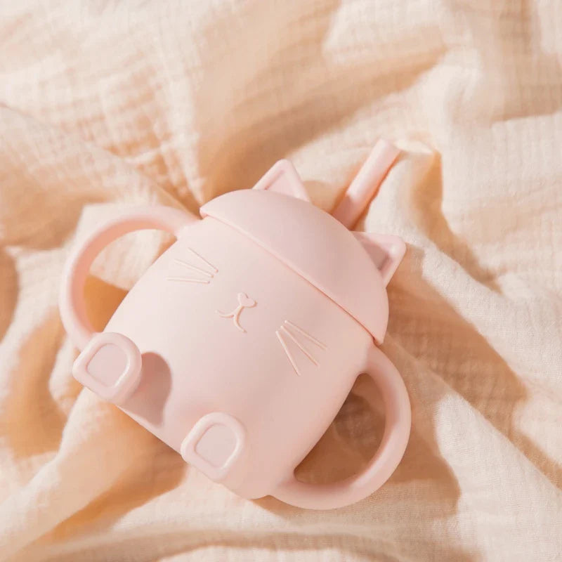 Der Hello World Trinkbecher Katze sitzend Rosa (13x11cm) ist ein rosafarbener Trinkbecher in Katzenform mit zwei Henkeln, Katzenohren auf dem Deckel und einem Strohhalm, der auf einem hellbeigen, strukturierten Stoff ruht.