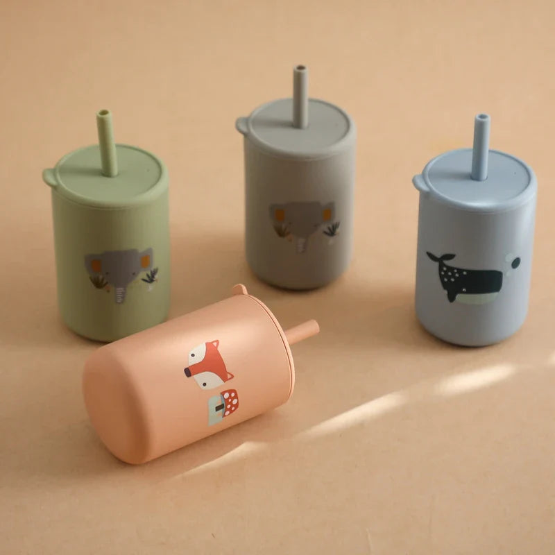 Trois gobelets Hello World verticaux avec un motif bleu (éléphant vert, éléphant gris, baleine bleue sans BPA) et un gobelet renard couleur pêche reposent sur un fond beige. Tous sont des gobelets de 6x12,5 cm aux couleurs pastel avec des motifs d'animaux et des pailles.