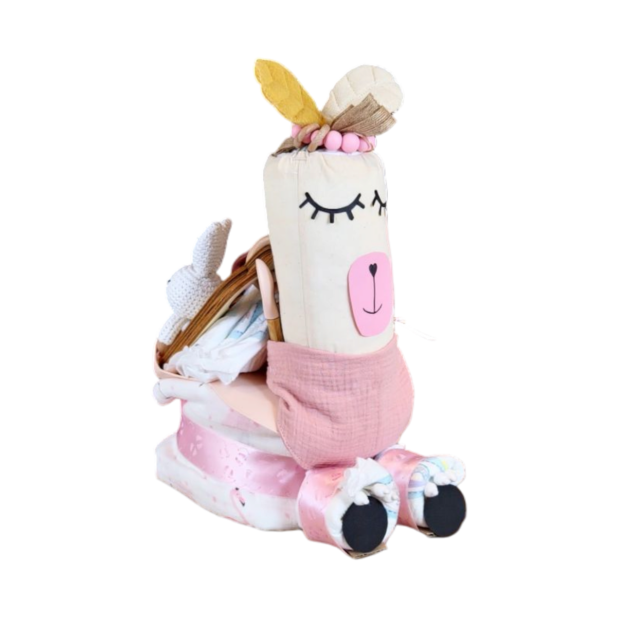 Das Hello World Windelgeschenk Lama (35x19cm), ein Plüschtier mit geschlossenen Augen, rosafarbenen Wangen und gelben Filzblättern, sitzt auf gefalteter rosa und weißer Babykleidung - ein bezauberndes Babygeschenkset mit kleinen Schuhen und Accessoires.