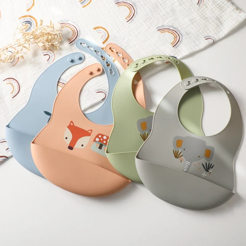 Vier wasserfeste Silikon-Babybibs von Hello World liegen auf dem Tisch: blau mit Bär, pfirsich mit Fuchs & Pilz, olivgrün mit Hase und der Latz mit Motiv Silikon Grau (30x21cm). Daneben eine weiße Stoffwindel mit Regenbogenmuster.