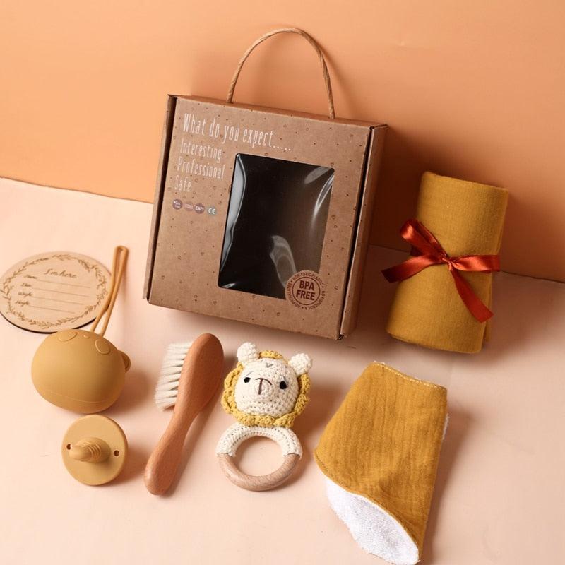 Das Leonie Baby Geschenk zur Geburt von Hello World enthält eine braune Box mit Klarsichtfenster, eine senfgelbe Decke, eine Notizkarte, eine Silikontrinkflasche, ein Schnuller-Set, eine Holzhaarbürste, einen Rassellöwe-Beißring und einen Waschlappen.