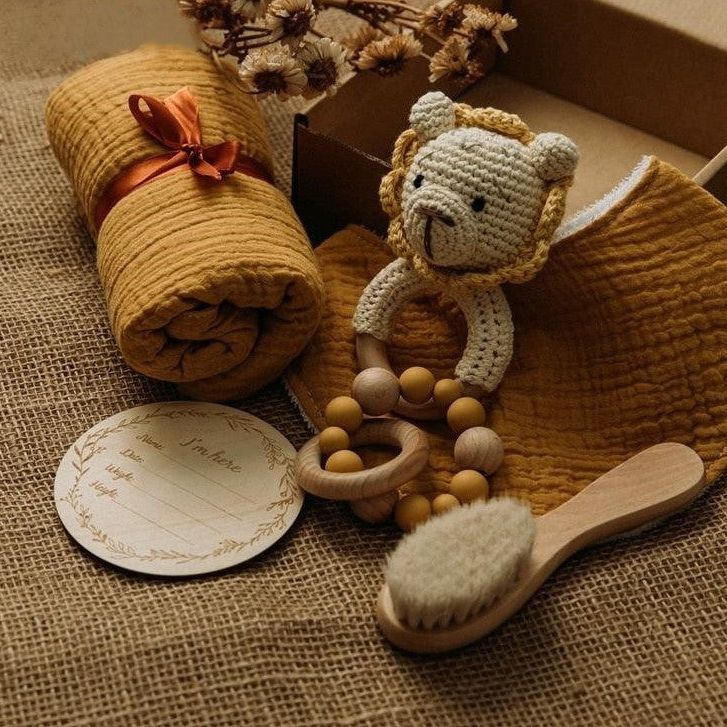 Das Lindor Baby Geschenk zur Geburt von Hello World enthält eine senfgelbe Decke, einen Beißring aus Holz Silikon, eine gehäkelte Löwenrassel, ein Lätzchen, Trockenblumen und eine runde Geschenkkarte - perfekt als Geschenk für ein Neugeborenes.