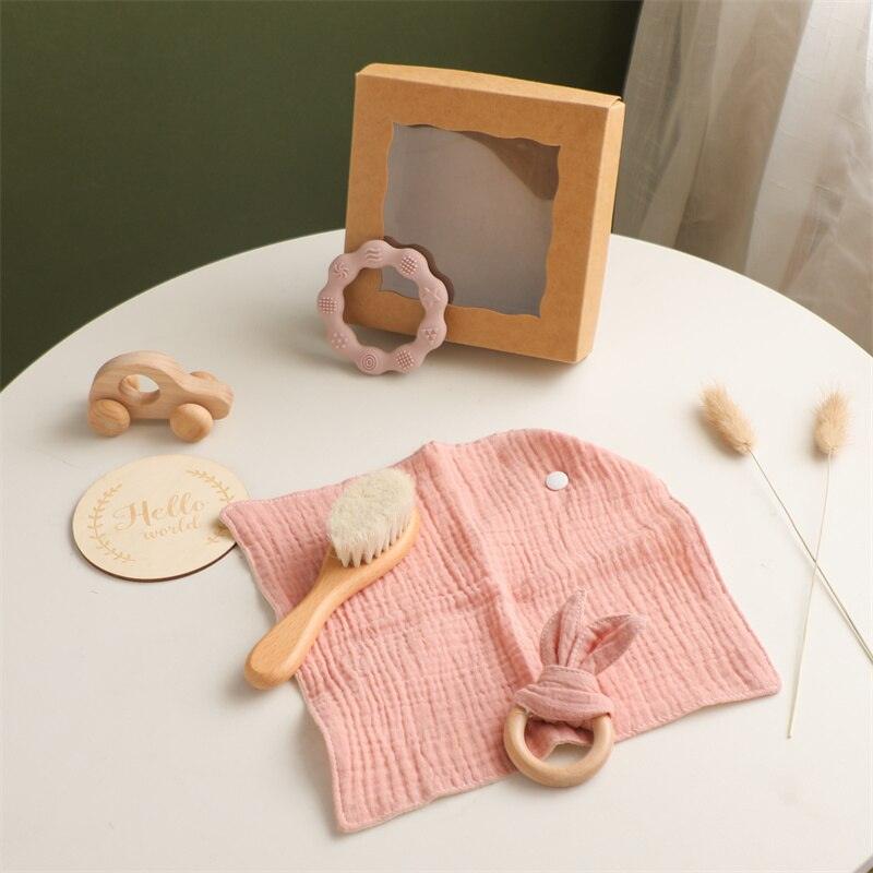 Auf einem runden Tisch steht das Lora Baby Geschenk zur Geburt von Hello World mit rosa Mulltuch, Holz-Greifling-Beißring, weicher Babyhaarbürste, Holzspielzeugauto, rosa Beißringen, "Hello World"-Schild und brauner Schachtel - charmant gestaltet mit getrockneten Gräsern.