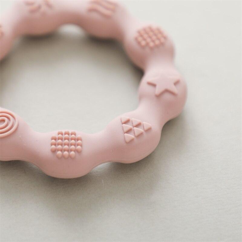 Eine Nahaufnahme des Lora Baby Geschenk zur Geburt von Hello World - ein zartrosa, kreisförmiger Beißring mit erhabenen Sternen, Dreiecken, Punkten und Wirbeln, der auf einer hellen Oberfläche ruht; ideal für Neugeborene.