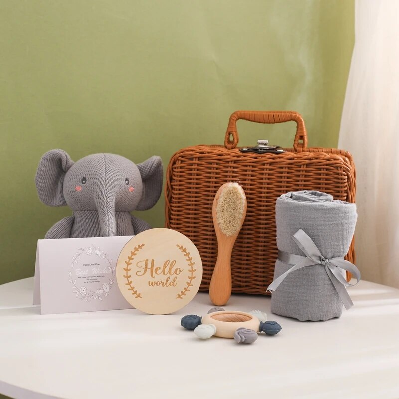Das Matteo Baby Geschenk zur Geburt von Hello World steht auf einem weißen Tisch mit einem grauen Plüschelefanten, einem Weidenkorb, einer Holzhaarbürste, einer gerollten Decke, einem Holz-Silikon-Beißring, Pastellsteinen, einer Grußkarte und einem Hello World Holzschild.