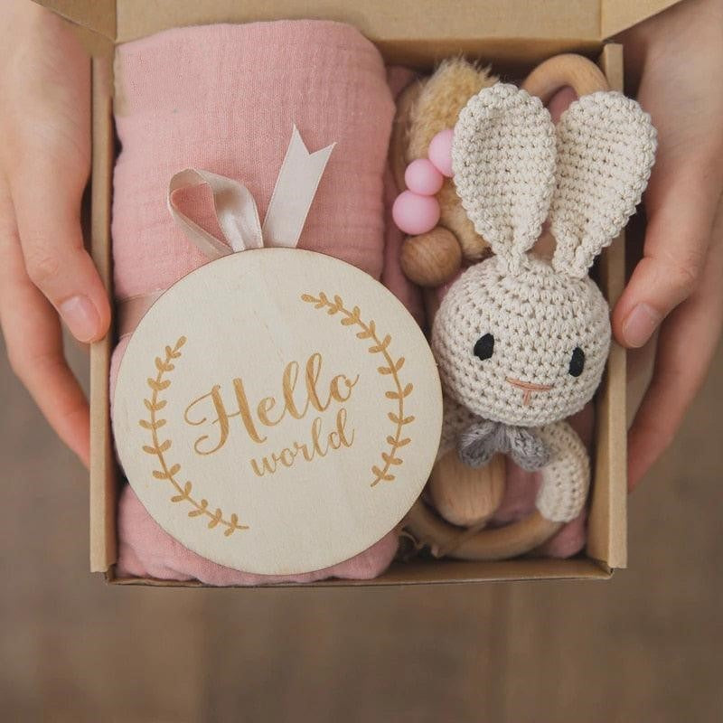Eine Person hält das Mia Baby Geschenk zur Geburt von Hello World in der Hand, das ein rosafarbenes Tuch, ein gehäkeltes Häschen, einen Beißring aus Holz-Silikon und eine "Hello world"-Holzplakette mit Blattrand enthält.