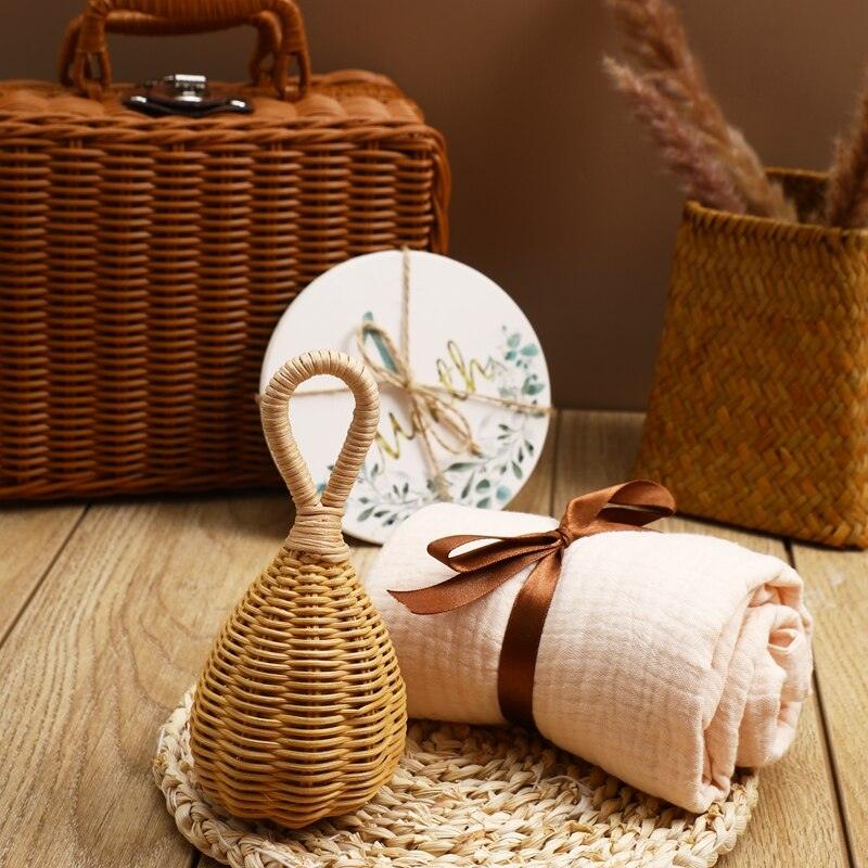 The Rusty Baby Newborn Gift from Hello World features a woven basket, a rolled blanket with a brown ribbon, a wicker rattle and a bird-themed plate on a wooden surface with a mat and a vase in the background.