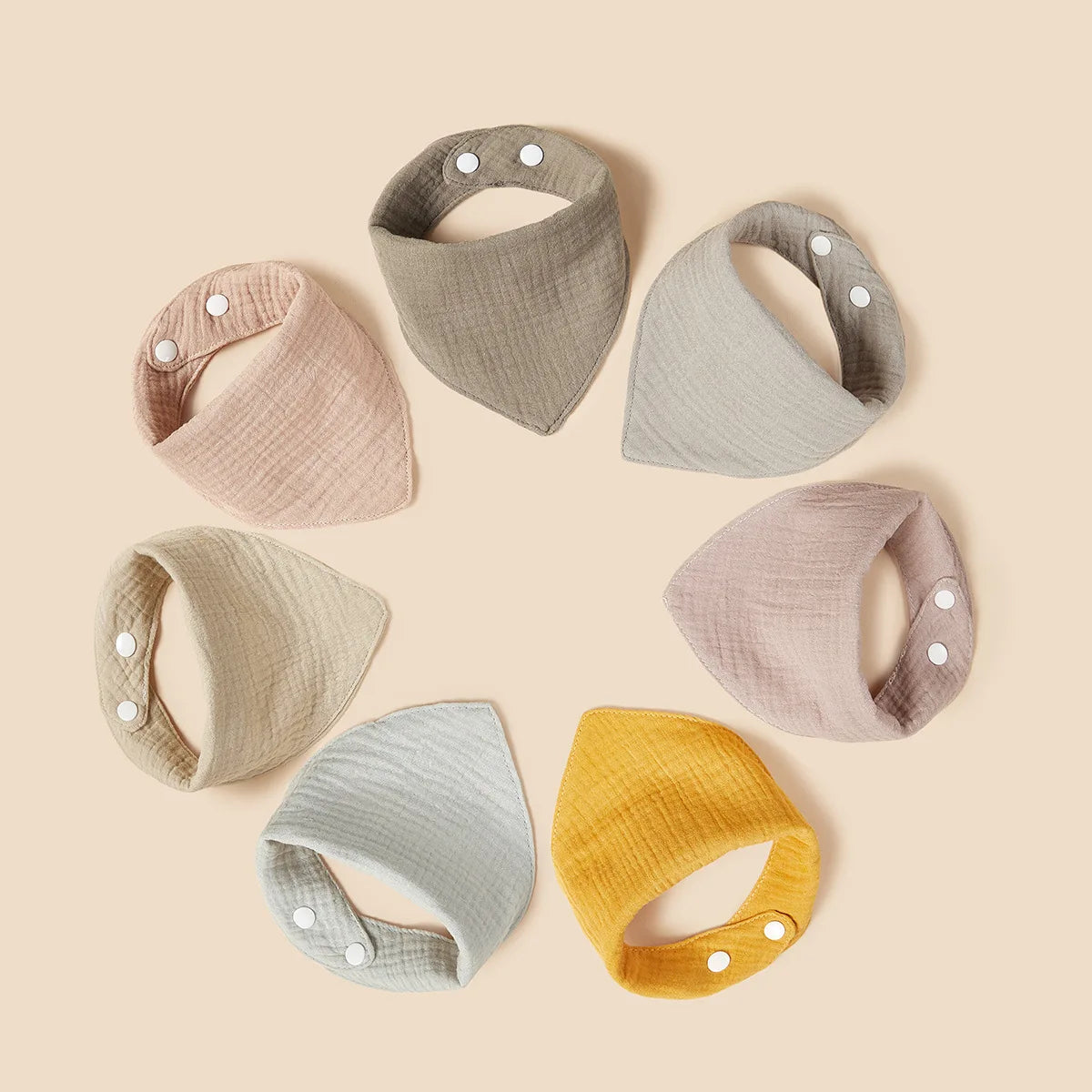 I sette bavaglini a bandana Hello World "Grey Cotton Bib" in toni pastello e neutri sono disposti in cerchio su uno sfondo beige e ciascuno ha due bottoni automatici bianchi su un angolo.