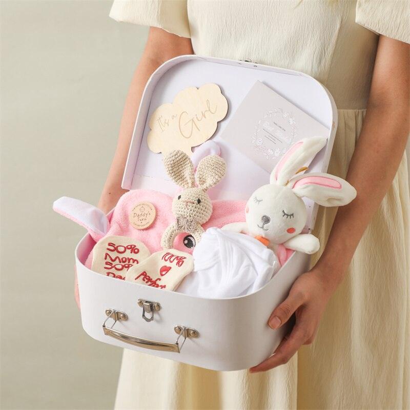 Eine Person in einem cremefarbenen Kleid hält einen offenen weißen Koffer mit Plüschhasen, Babykleidung und Karten - Selma Baby Geschenk zur Geburt von Hello World, dem perfekten Mädchen Geschenkset und Prinzessinnen Ausstattung.