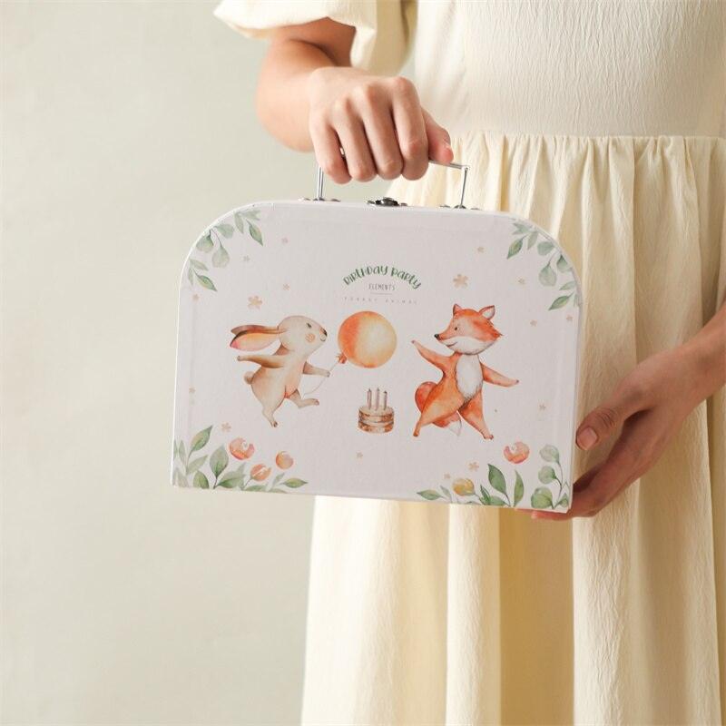 Eine Person in einem cremefarbenen Kleid hält das Hello World Selma Baby Geschenk zur Geburt, einen weißen Koffer mit aquarellierten Hasen und Füchsen in der Nähe einer Geburtstagstorte, perfekt als ein Mädchen Geschenkset und mit floralen Akzenten verziert.