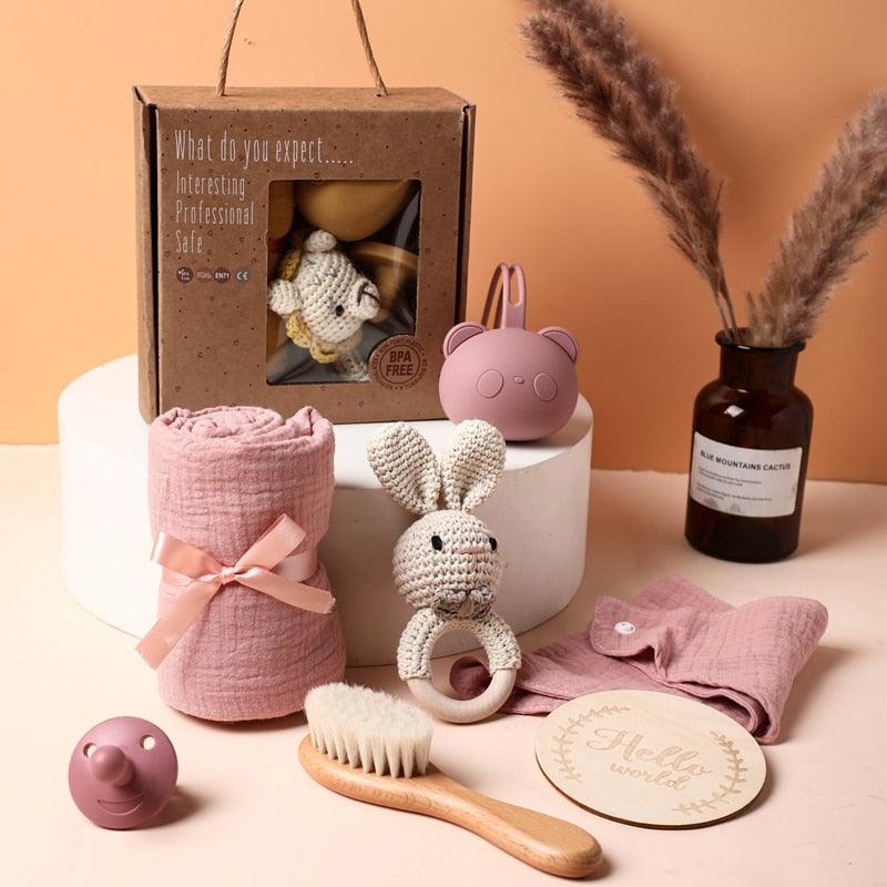 Das Serena Baby Geschenk zur Geburt von Hello World besteht aus einem gehäkelten Rasselhasen, einer rosa Musselindecke, einer Holzbürste, einer runden Plakette, einem Schnuller-Set (rosa Schnuller & Beißring) und einer braunen Vase mit Pampasgras.