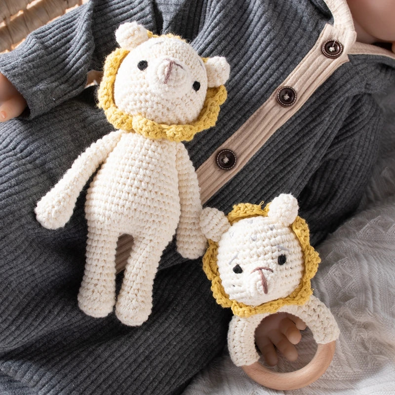 Il peluche leone lavorato a maglia Hello World (18,5 x 10 cm) e il leone abbinato con la criniera gialla su un anello di legno stanno accanto a un bambino in una tutina grigia con finiture marrone chiaro.