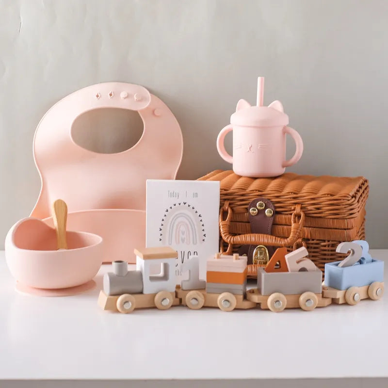 Das Tiffany Baby Geschenk zur Geburt von Hello World besteht aus einem pastellfarbenen Fütterungsset, einem Weidenkorb, einer Karte und einer Holz-Lokomotive mit Nummern - charmant und nostalgisch auf einer weißen Oberfläche vor einem hellen Hintergrund.