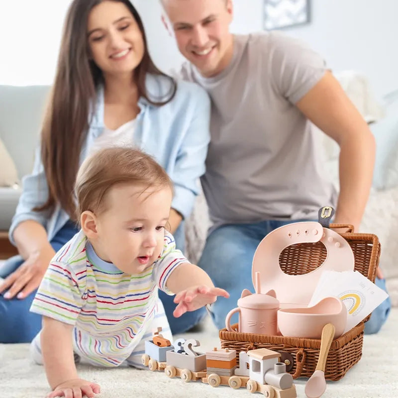 Ein Baby krabbelt auf einem Teppichboden und greift nach dem Hello World Tiffany Baby Geschenk zur Geburt, während zwei Erwachsene im Hintergrund lächeln. In der Nähe steht ein Korb mit Babyartikeln, die Erinnerungen an die Abenteuer der frühen Kindheit wachrufen.