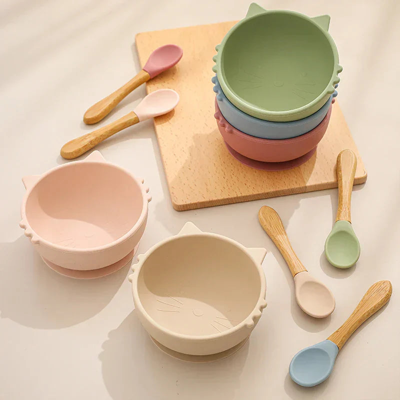 Vier pastellfarbene katzenförmige Schalen und fünf Löffel mit Holzgriff sind auf einer hellen Fläche angeordnet, mit einem Holzbrett im Hintergrund. Die Schalen sind in Rosa, Beige, Grün und Blau gehalten.