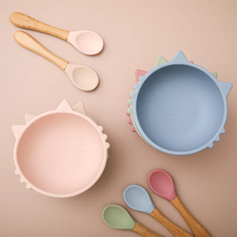 Zwei katzenförmige Schalen, eine rosa und eine blaue, stehen auf einer beigen Fläche, daneben vier Silikonlöffel mit Holzgriff in Pastellfarben.