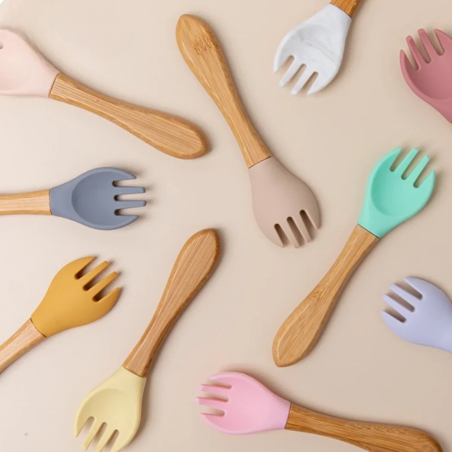 Eine flache Reihe von Babylöffeln mit Holzgriff und bunten Silikonspitzen in den Farben Rosa, Gelb, Grau, Grün, Weiß, Hellbraun, Lila und Blau, angeordnet auf einem beigen Hintergrund.